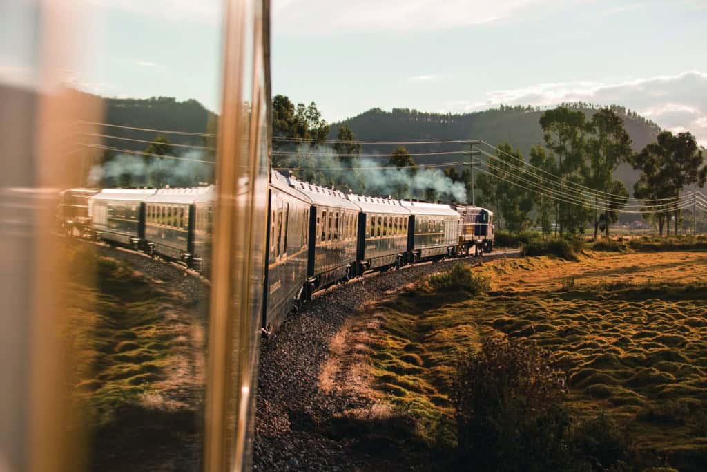 PeruRail train traveling through a rural landscape with hills, green pastures, and patches of grass, set against the Andes mountains.