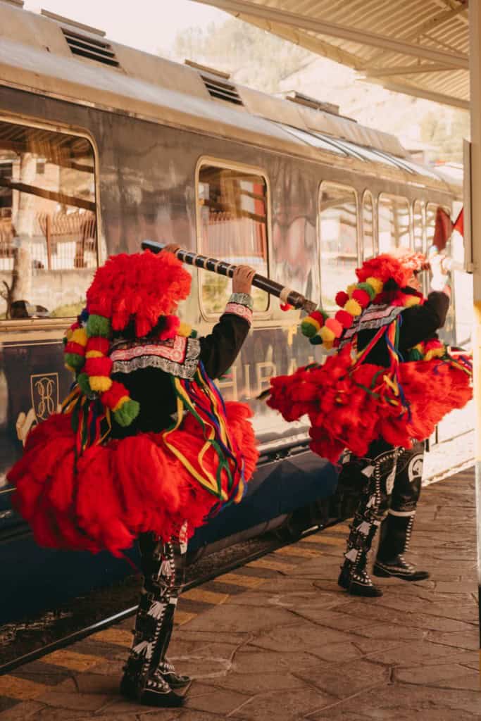 Traditional Peruvian dancers in vibrant red costumes and colorful accessories performing near a PeruRail train at a station.