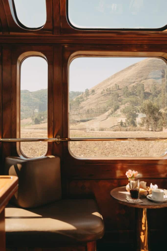 Elegant train interior featuring a wooden frame and large windows offering a scenic view of rolling hills and fields, with a table set for refreshments, including coffee and flowers.