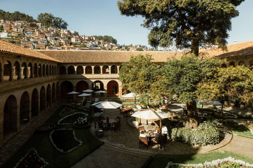 Colonial courtyard of a historic building with arched walkways, manicured gardens, and outdoor dining tables shaded by umbrellas under a large tree, with a hillside cityscape in the background.