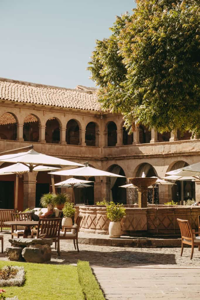 Close-up of a serene colonial courtyard featuring a central stone fountain, lush greenery, and outdoor seating with white umbrellas, surrounded by arched stone architecture and warm sunlight at a Peru hotel.