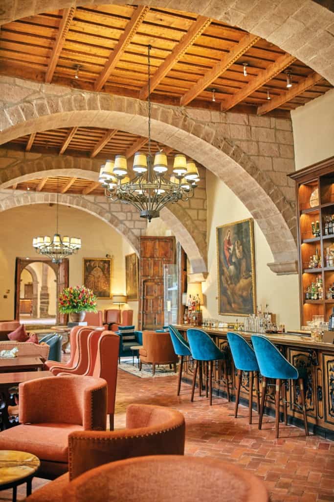 Elegant lounge and bar area with warm terracotta tones, velvet blue bar stools, and plush seating, featuring arched stone ceilings, rustic wooden beams, chandeliers, and classic artwork for a luxurious ambiance at a Peru Hotel.