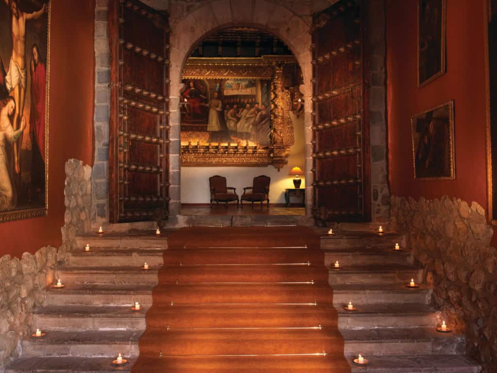 Elegant interior of a historic building featuring a grand entrance with open wooden doors, stone walls, candle-lit steps, and an ornate golden altar in the background at a Peru Hotel.