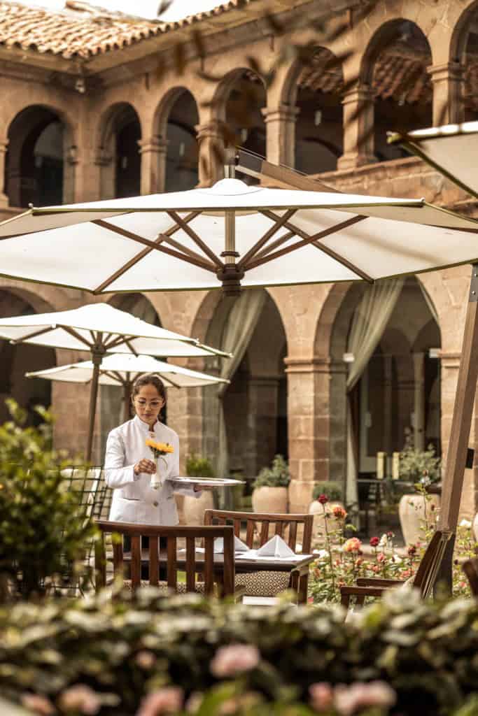 Woman serving drinks while working in colonial courtyard of a Peru hotel with pristine tables, green gardens, and flowers in the background.